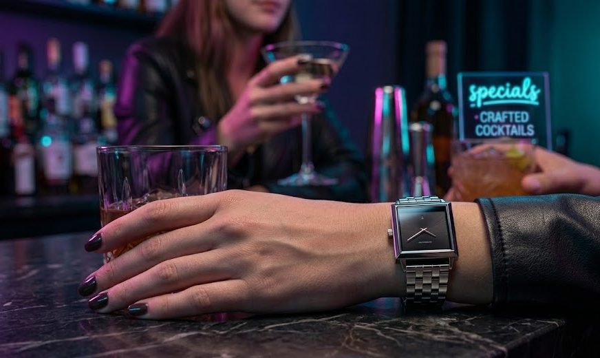 Mulher com jaqueta de couro em um lounge noturno segurando um drink, com foco no pulso usando um moderno relógio Guess quadrado de fundo preto.