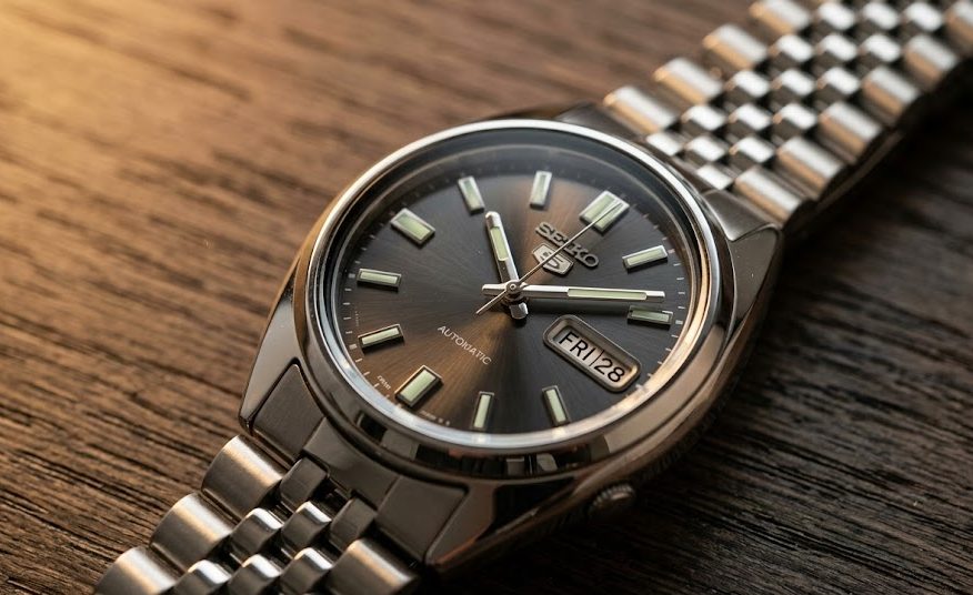 Fotografia em close-up de um elegante relógio Seiko 5 Automático mecânico de aço inoxidável com mostrador cinza escuro luminoso, semelhante ao Seiko SNXS79, sobre uma mesa de carvalho.