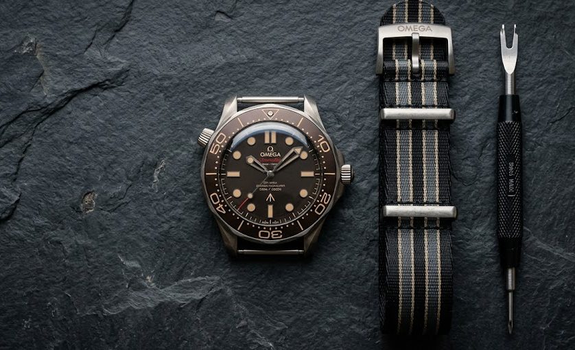 Relógio Omega Seamaster Relógio Omega Seamaster: Pulseiras estilo NATO nas cores do James Bond (preto e cinza) ao lado de um relógio de mergulho e uma ferramenta de troca de pinos.